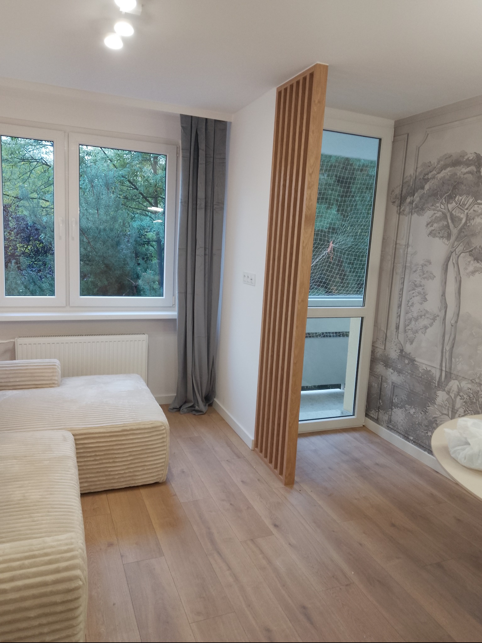 Jasne wnętrze po wykończeniu: drewniana podłoga, sofa, okno z widokiem na zieleń, dekoracyjna ścianka z drewnianych listew i tapeta w stylu retro z motywem drzew.