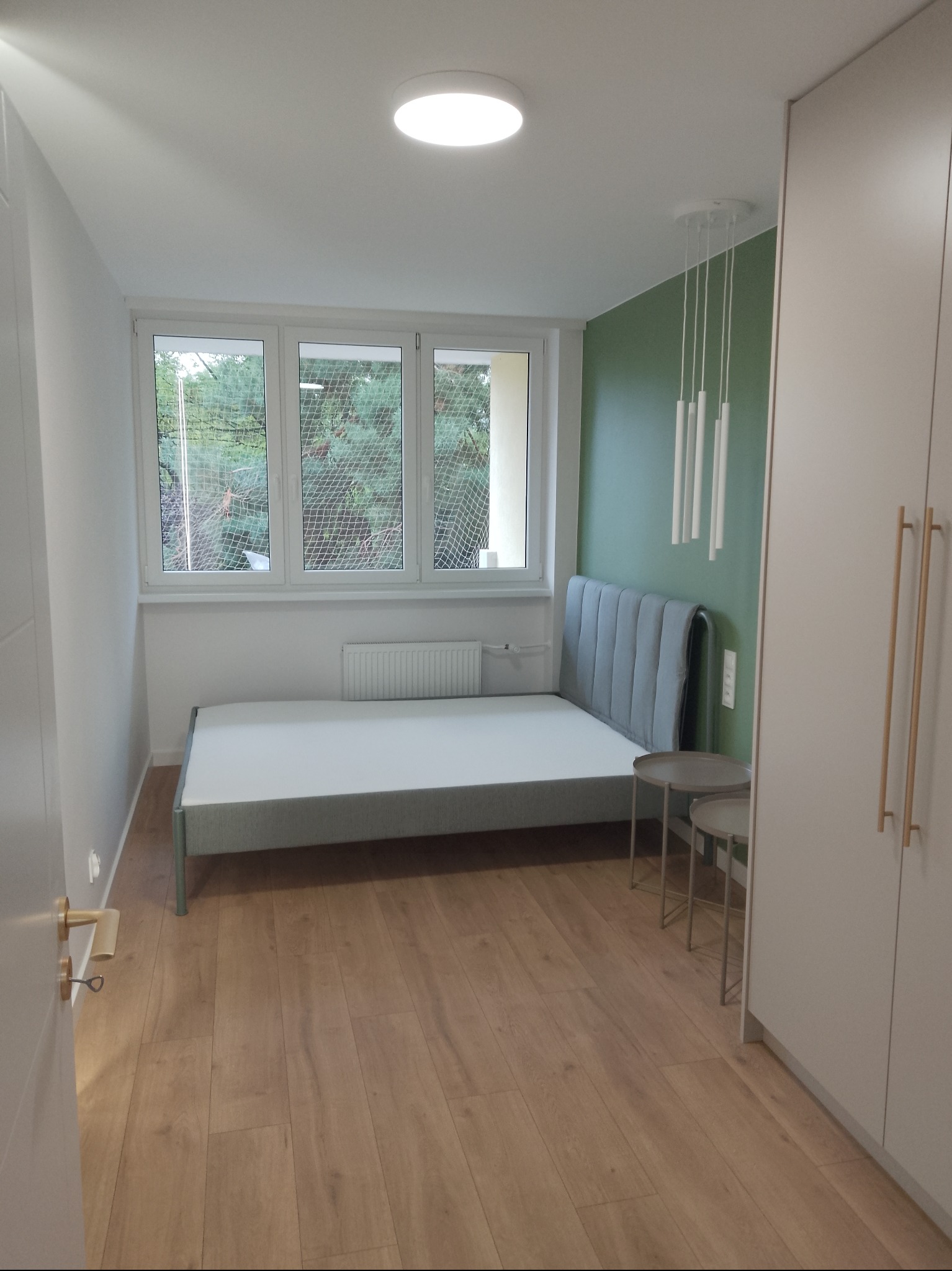 Jasne wnętrze sypialni po remoncie: drewniana podłoga, szara rama łóżka z zagłówkiem, białe ściany i szafa, akcentująca zielona ściana za łóżkiem, okno z widokiem na drzewa.