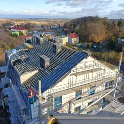 Artur Jaśkiewicz Kierownik Budowy - Wysoki kąt widzenia na budynek w trakcie budowy dachu z widocznymi rusztowaniami, elementami konstrukcji dachowej i kominami z logo producenta, na tle zabudowy mieszkalnej i krajobrazu.