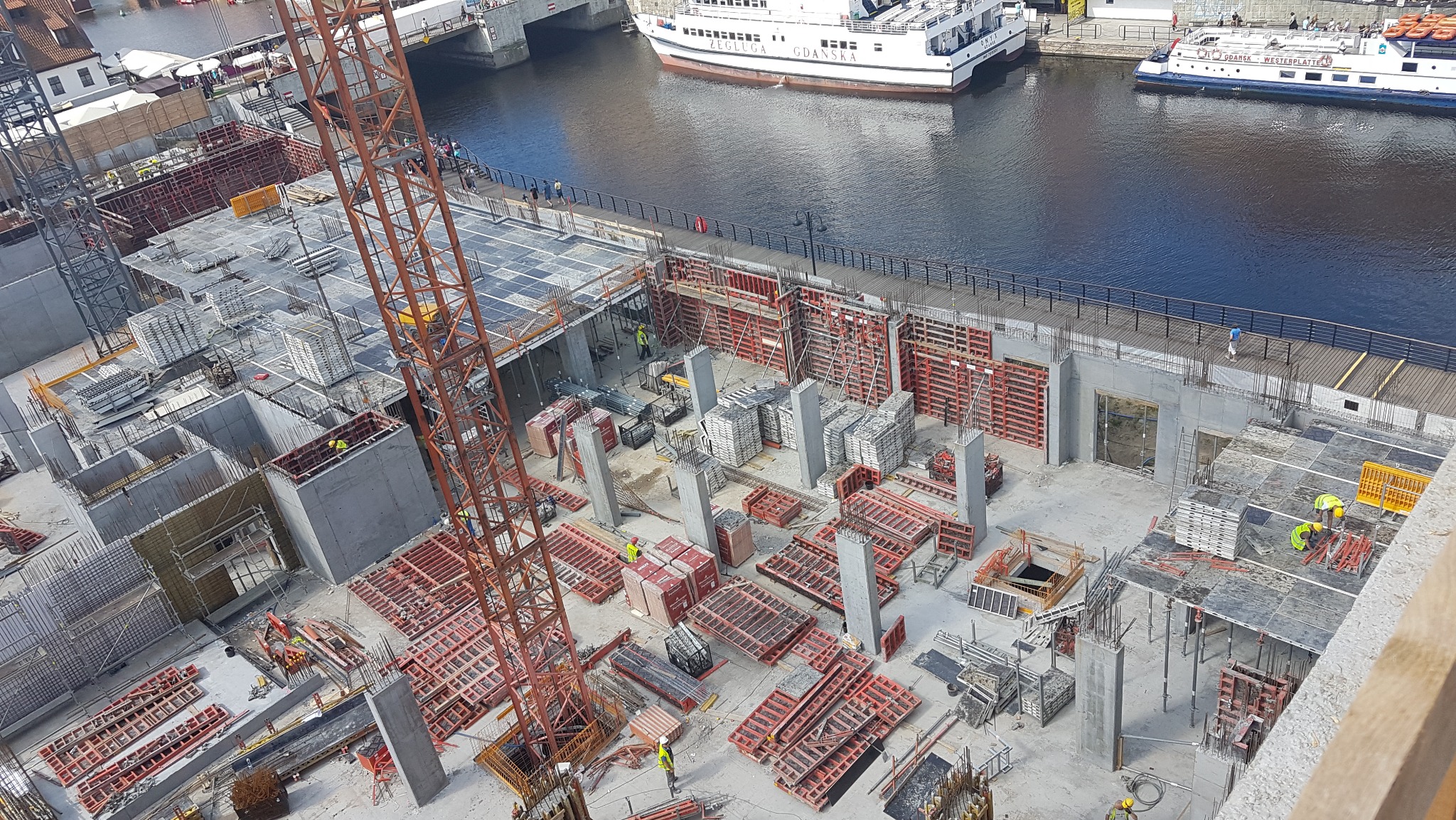 Widok z góry na plac budowy w Gdańsku, z widocznymi żurawiem, zbrojeniami, szalunkami, betonowymi słupami i fragmentem rzeki Motławy z pływającymi statkami pasażerskimi w tle.