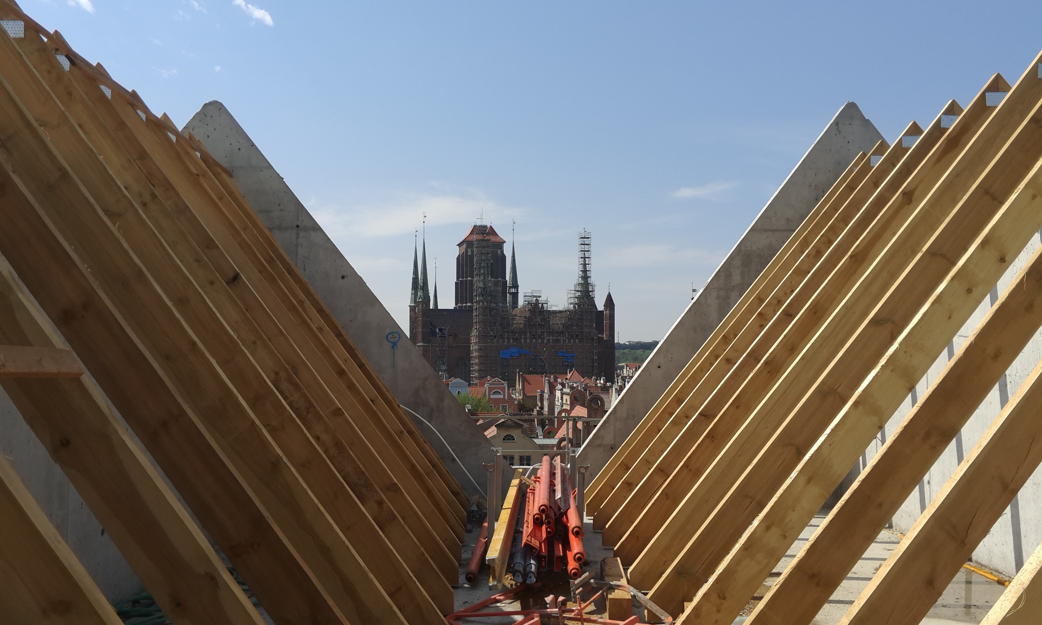Konstrukcja dachu z drewnianych krokwi i betonowych ścian szczytowych, w tle widok na gotycką bazylikę Mariacką w Gdańsku w trakcie renowacji, częściowo osłoniętą rusztowaniami.