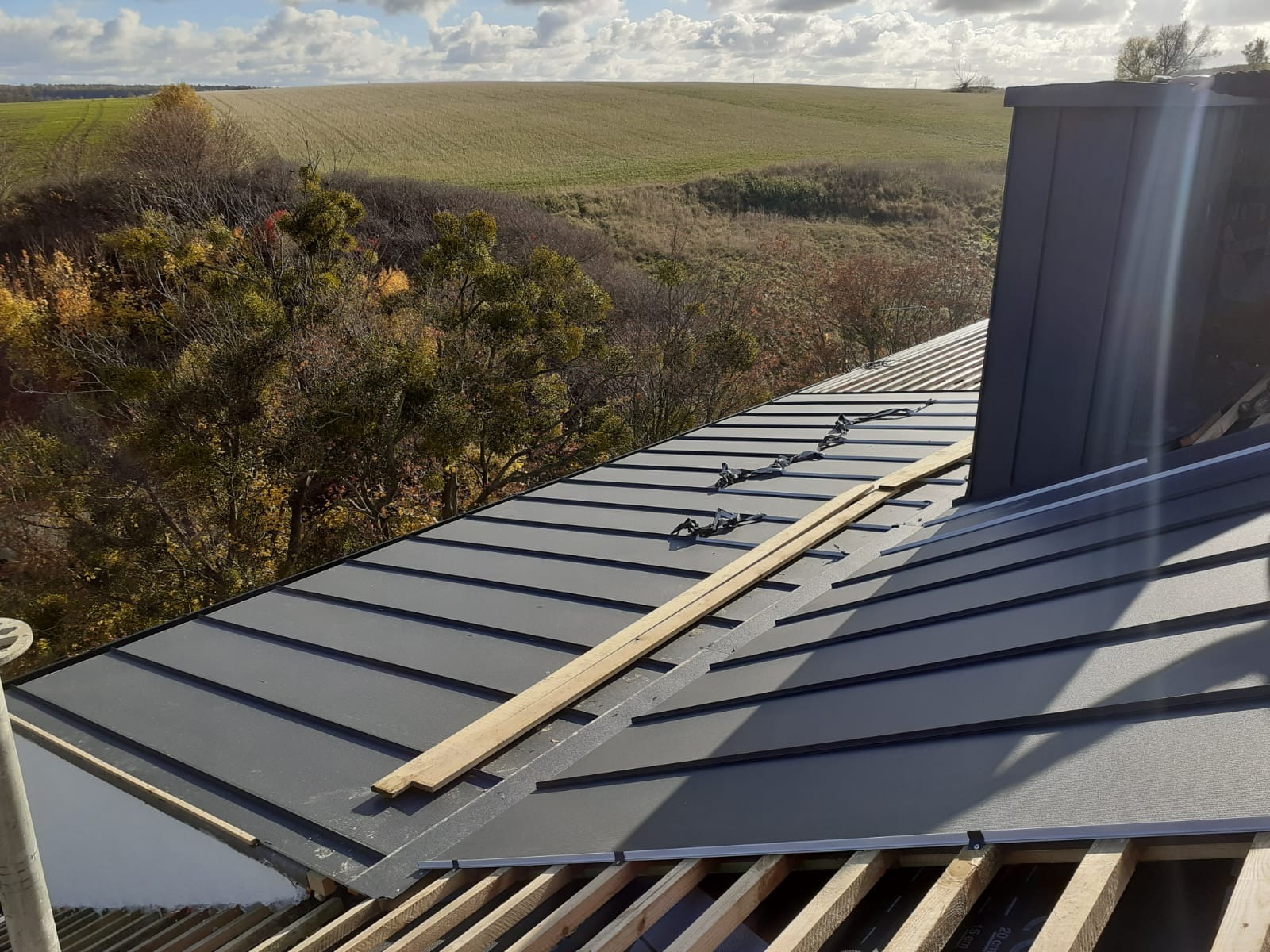 Montaż blachy na dachu z widokiem na jesienny krajobraz, drewniane łaty i elementy mocujące widoczne na powierzchni blachy.