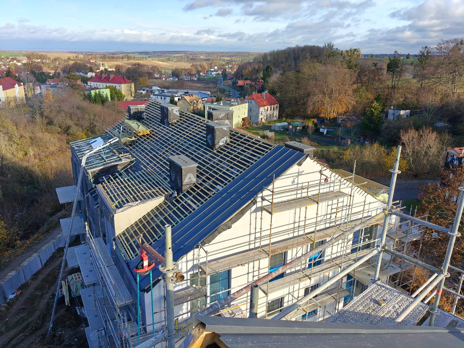 Wysoki kąt widzenia na budynek w trakcie budowy dachu z widocznymi rusztowaniami, elementami konstrukcji dachowej i kominami z logo producenta, na tle zabudowy mieszkalnej i krajobrazu.