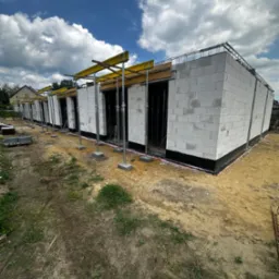 Wznoszenie parterowego budynku z bloczków, widoczne deskowanie i zbrojenie wieńca. Budowa w toku, słoneczny dzień.