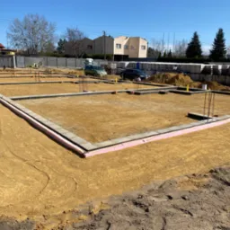 Fundamenty domu jednorodzinnego z izolacją termiczną, przygotowane do dalszych prac budowlanych. Widoczne zbrojenia i palety z bloczkami fundamentowymi.