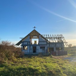 VITA-BUD - Wznoszony dom jednorodzinny z białych bloczków, z częściowo wykonaną konstrukcją dachu. W tle błękitne niebo i zielona trawa. Widoczne palety z cegłami.