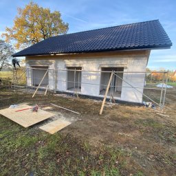 VITA-BUD - Wznoszony dom z białych bloczków, z czarnym dachem i otworami okiennymi, otoczony rusztowaniami. Na pierwszym planie leżą płyty i narzędzia.