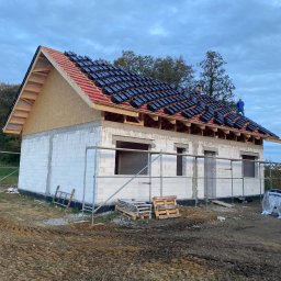 VITA-BUD - Budowa domu z białych bloczków: ściany wzniesione, dach w trakcie krycia czarną dachówką. Widoczni pracownicy na dachu, rusztowania i palety na błotnistym terenie.