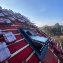 Montaż okna dachowego na czerwonym dachu pokrytym częściowo dachówką, widok z góry w słoneczny dzień. Dachówka ułożona w stosach, zabezpieczona gumkami.