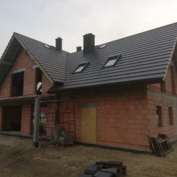 VITA-BUD - Niedokończony dom z czerwonej cegły z ciemnym dachem i oknami dachowymi, w tle pochmurne niebo i drzewa.