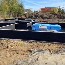 VITA-BUD - Fundamenty domu w trakcie budowy, zabezpieczone czarną izolacją, z widocznymi materiałami budowlanymi i ziemią w tle, w słoneczny dzień.