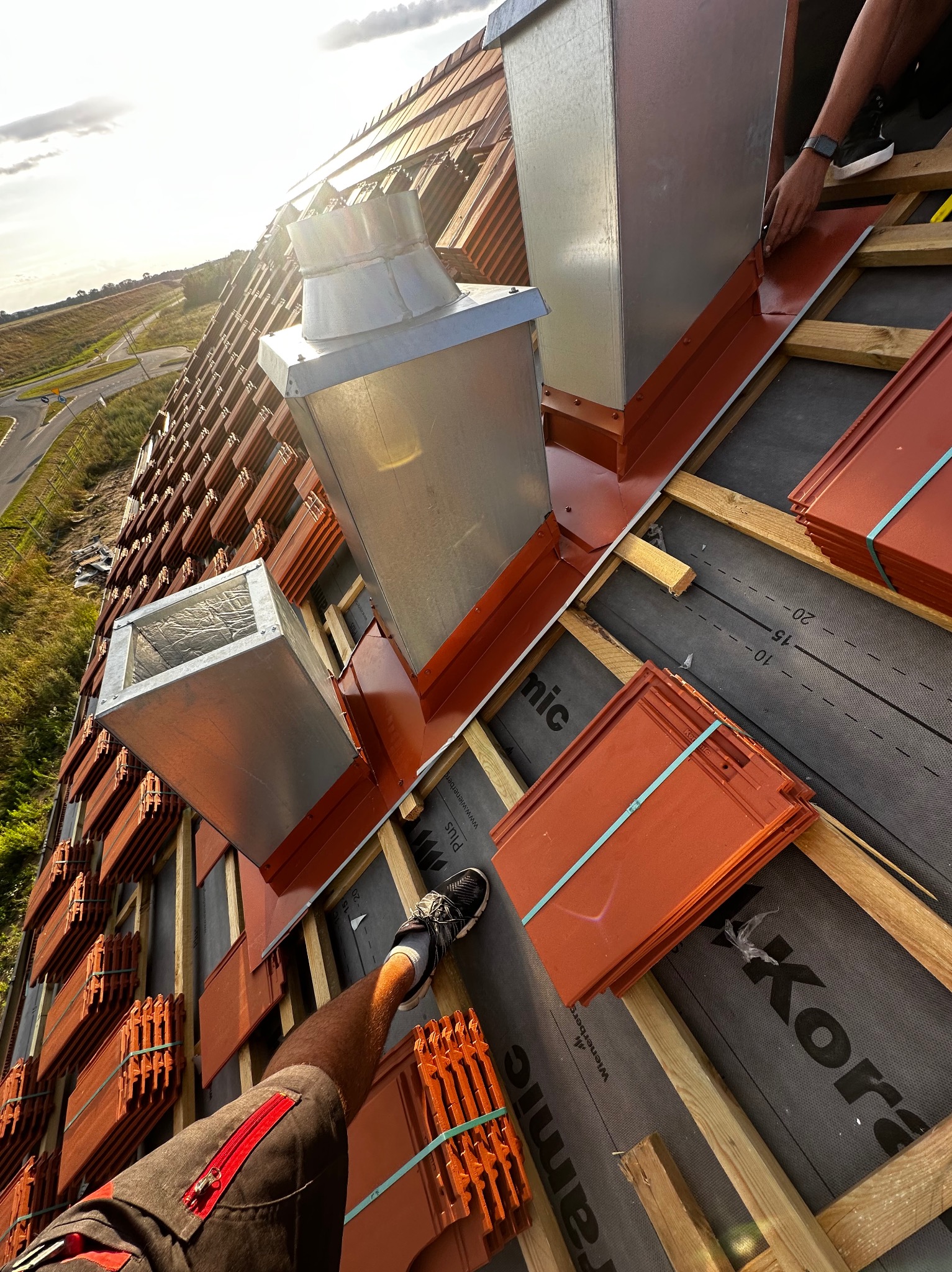 Montaż blaszanych kominów wentylacyjnych i dachówki ceramicznej na drewnianej konstrukcji dachu, widok z góry na częściowo ukończony dach.