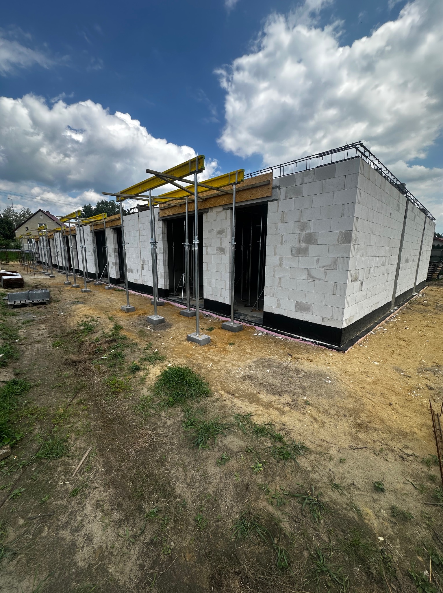 Wznoszenie parterowego budynku z bloczków, widoczne deskowanie i zbrojenie wieńca. Budowa w toku, słoneczny dzień.