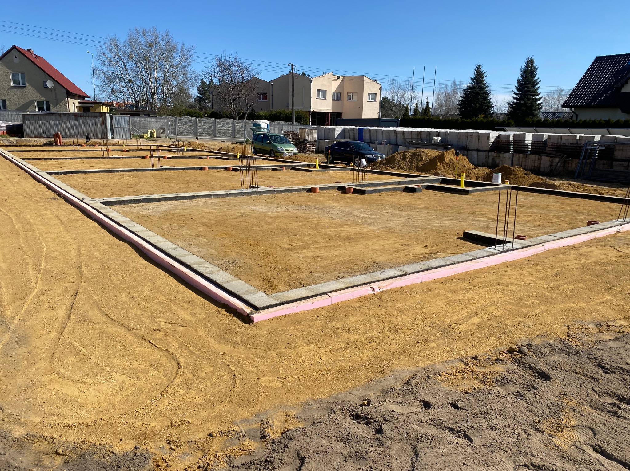 Fundamenty domu jednorodzinnego z izolacją termiczną, przygotowane do dalszych prac budowlanych. Widoczne zbrojenia i palety z bloczkami fundamentowymi.