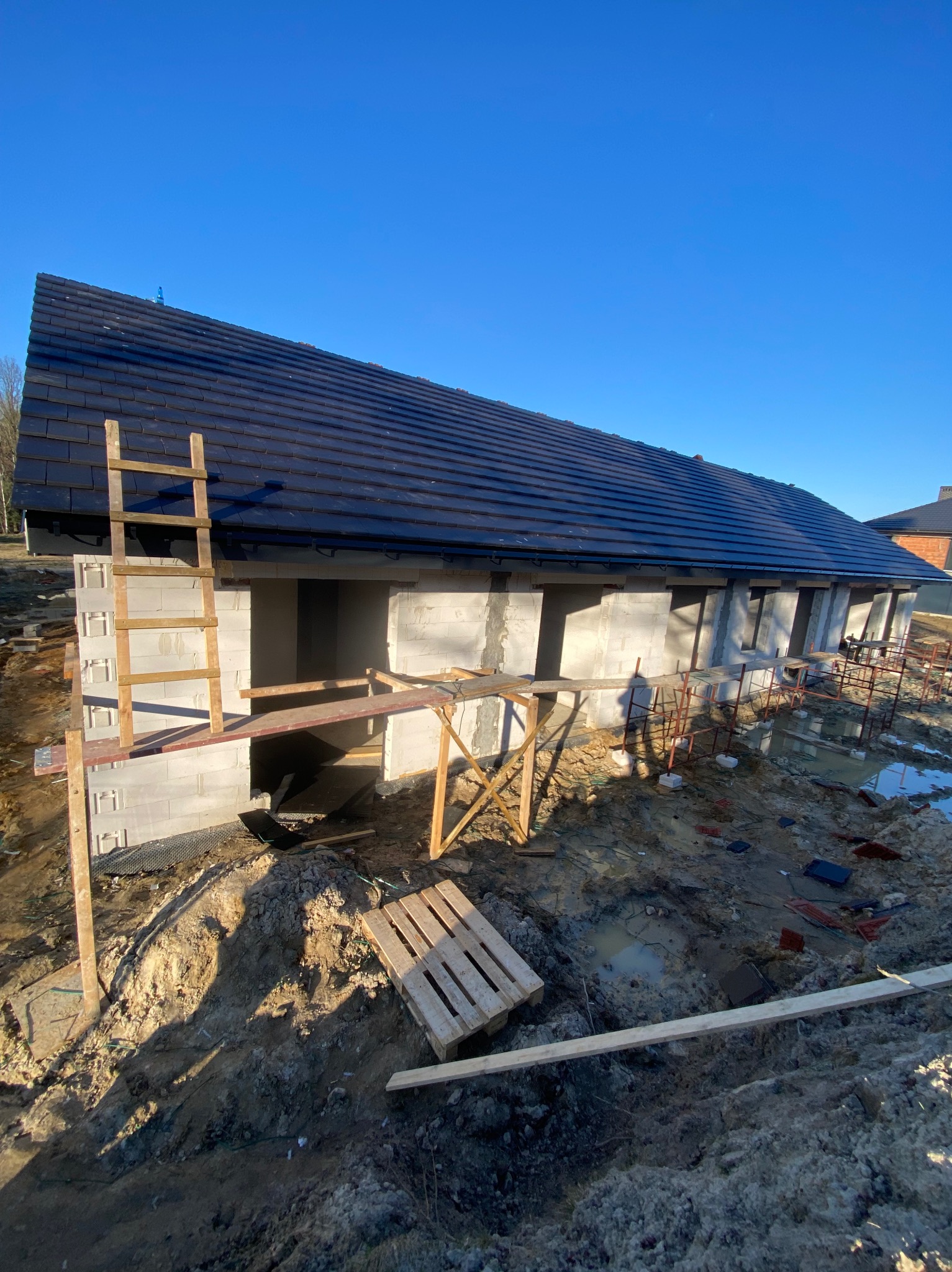 Dom w budowie: ściany z bloczków, czarny dach z dachówki, drewniana drabina i rusztowania. Wokół błoto i plac budowy w słoneczny dzień.
