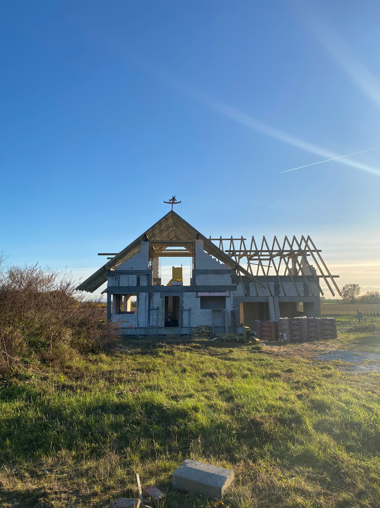 Wznoszony dom jednorodzinny z białych bloczków, z częściowo wykonaną konstrukcją dachu. W tle błękitne niebo i zielona trawa. Widoczne palety z cegłami.