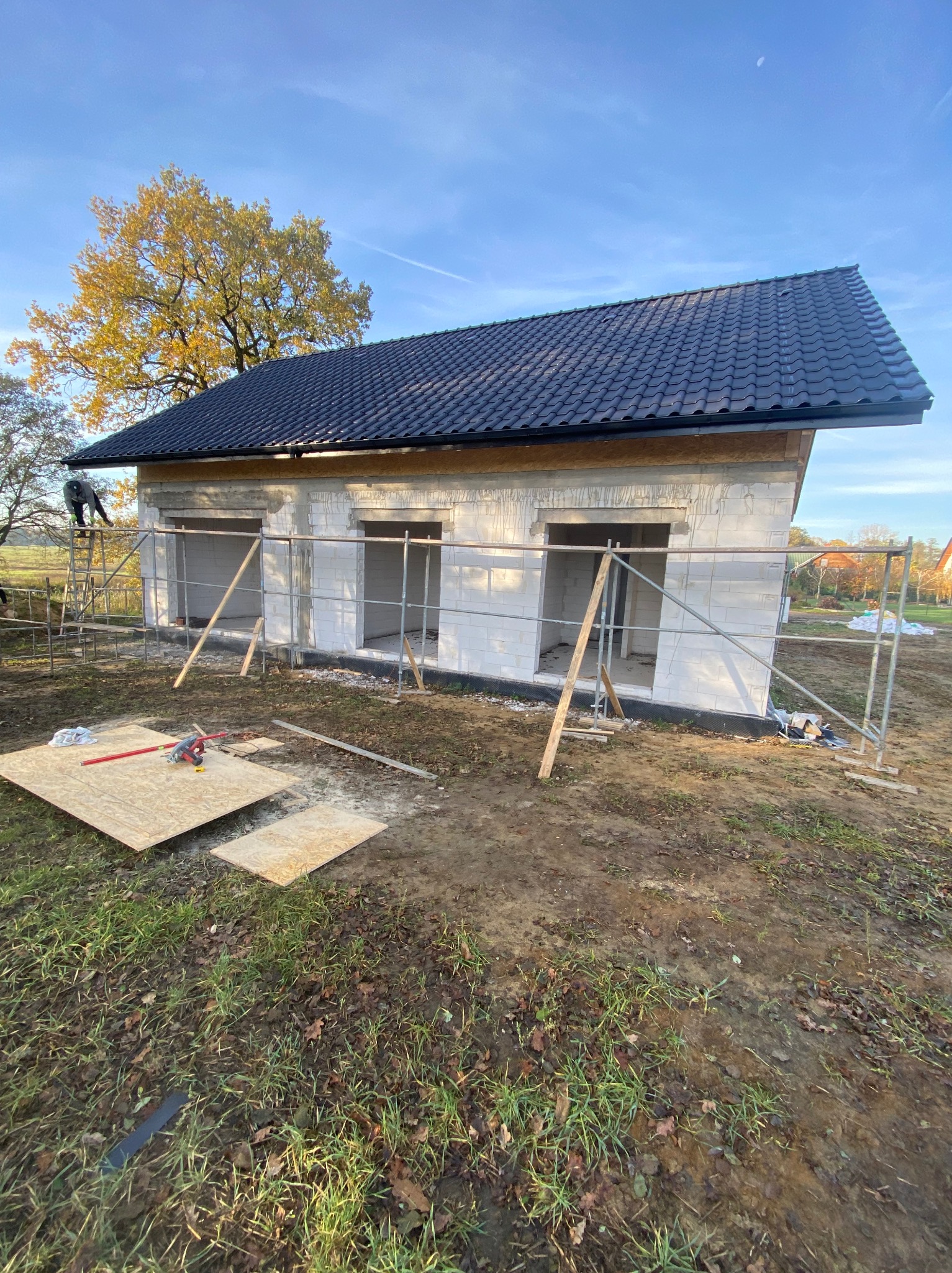 Wznoszony dom z białych bloczków, z czarnym dachem i otworami okiennymi, otoczony rusztowaniami. Na pierwszym planie leżą płyty i narzędzia.