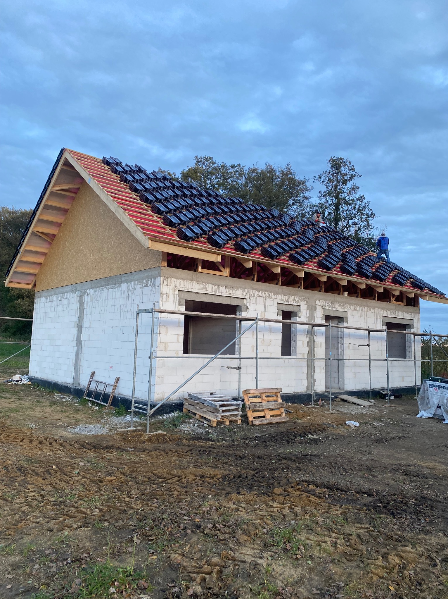 Budowa domu z białych bloczków: ściany wzniesione, dach w trakcie krycia czarną dachówką. Widoczni pracownicy na dachu, rusztowania i palety na błotnistym terenie.
