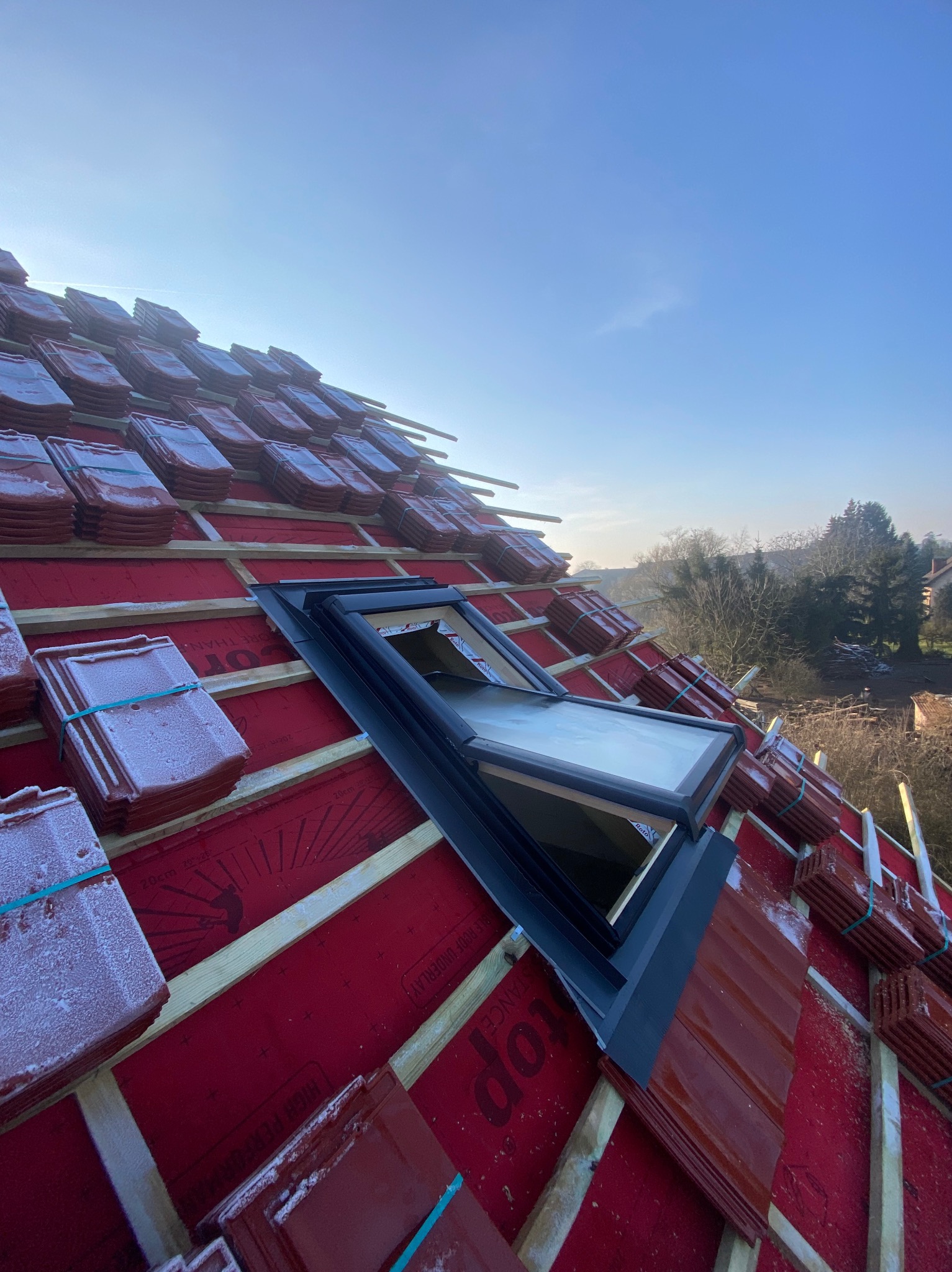 Montaż okna dachowego na czerwonym dachu pokrytym częściowo dachówką, widok z góry w słoneczny dzień. Dachówka ułożona w stosach, zabezpieczona gumkami.