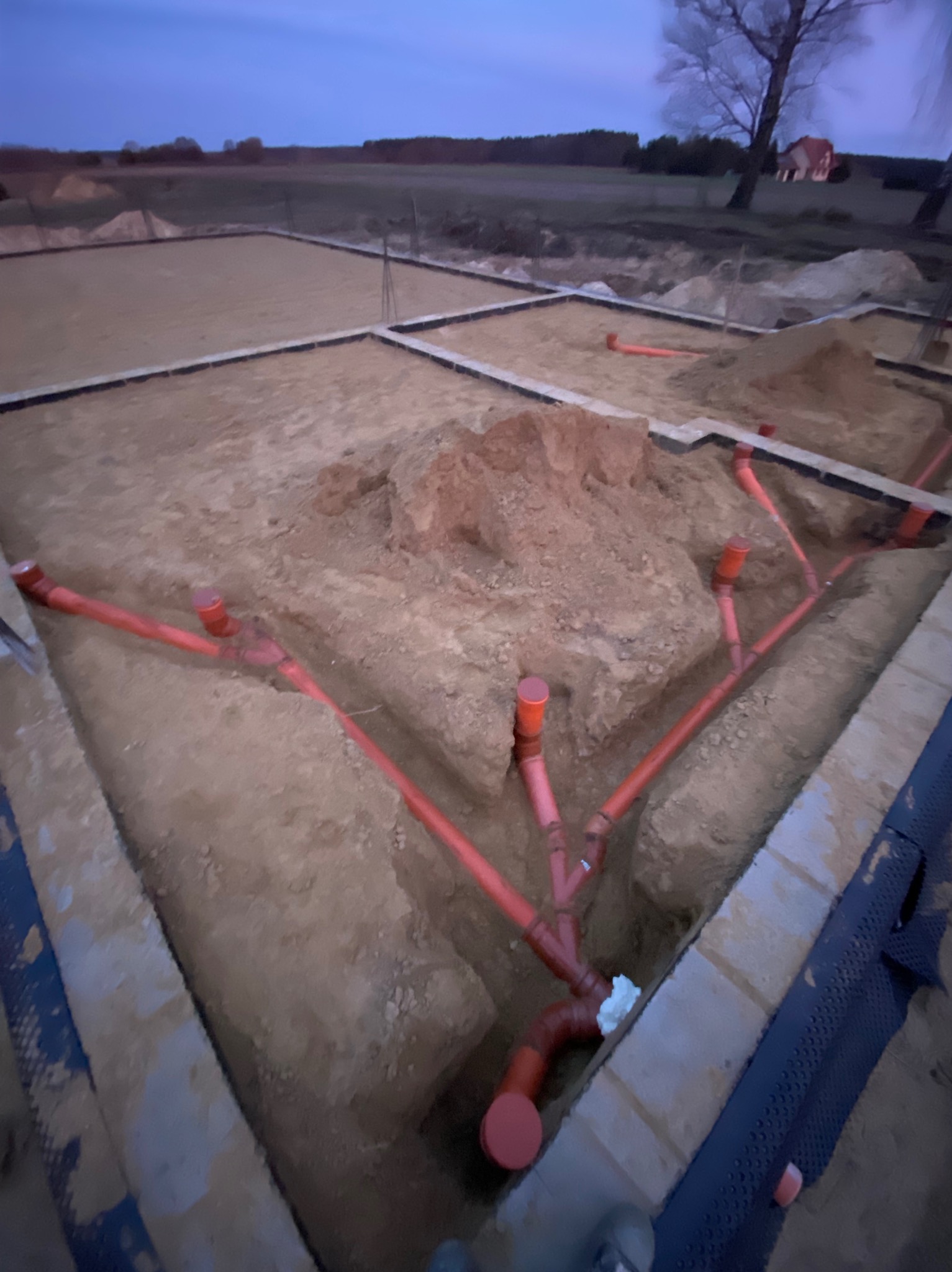 Fundamenty domu w trakcie budowy: widoczne szalunki, wykop i instalacja kanalizacyjna z pomarańczowych rur. W tle krajobraz polny pod wieczornym niebem.
