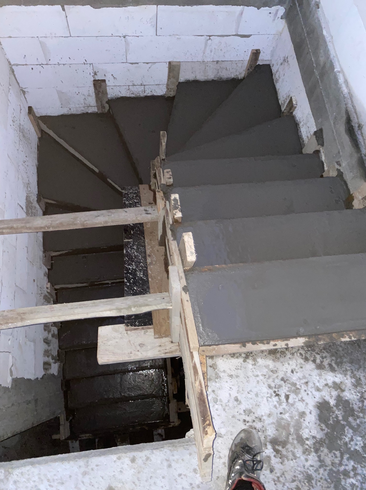 Betonowe schody spiralne w trakcie budowy, widok z góry, z drewnianym szalunkiem i fragmentem buta w rogu. Surowe wnętrze budynku z białymi bloczkami.