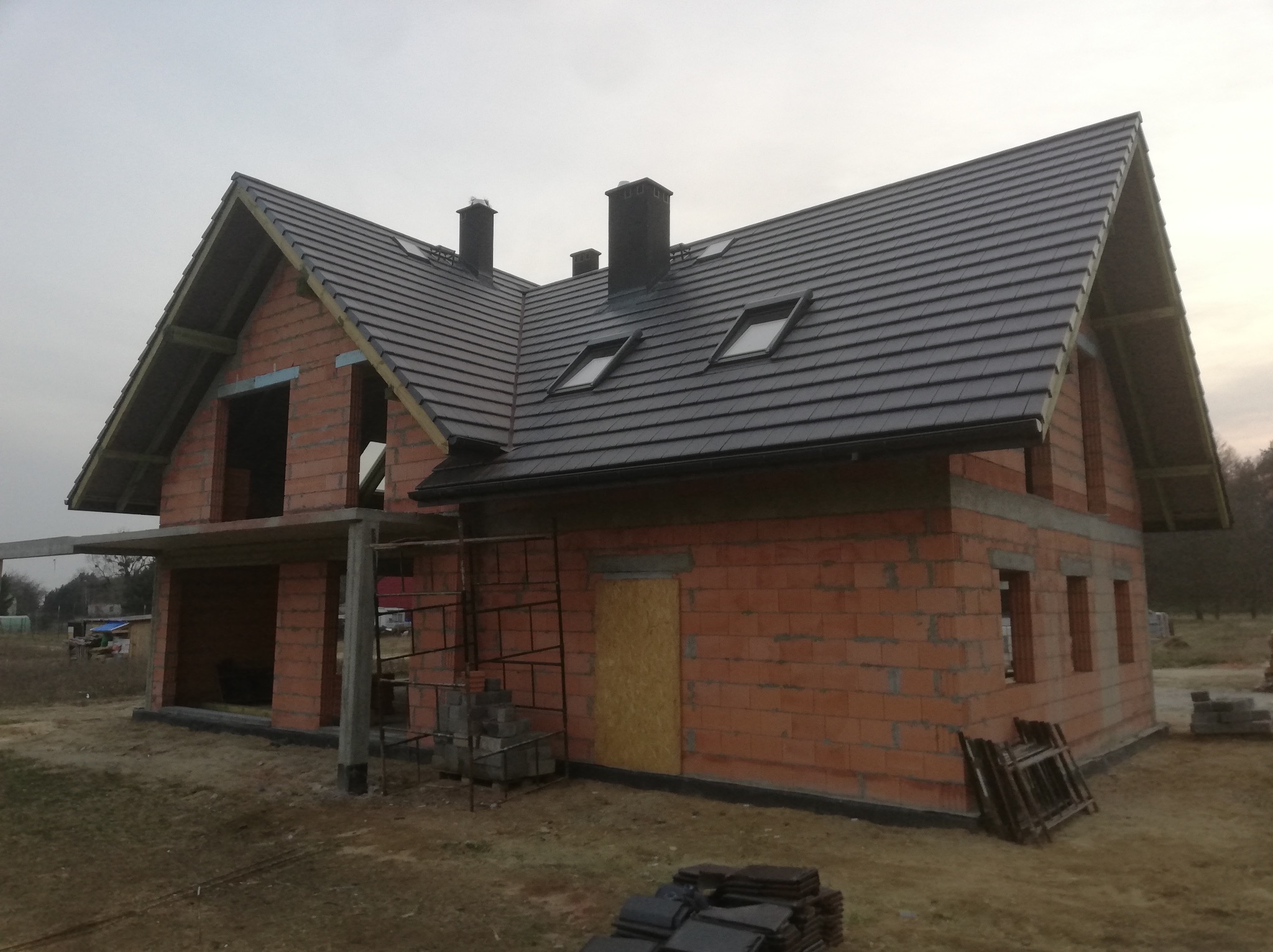 Niedokończony dom z czerwonej cegły z ciemnym dachem i oknami dachowymi, w tle pochmurne niebo i drzewa.