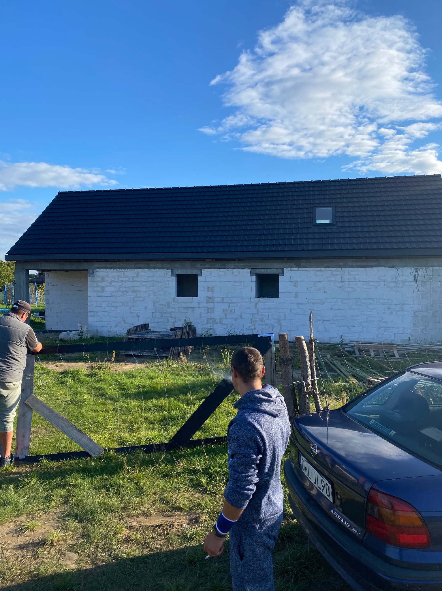 Budynek w stanie surowym zamkniętym z ciemnym dachem, białe ściany z pustaków, widoczne okna dachowe i otwory okienne. Na pierwszym planie osoby i samochód.