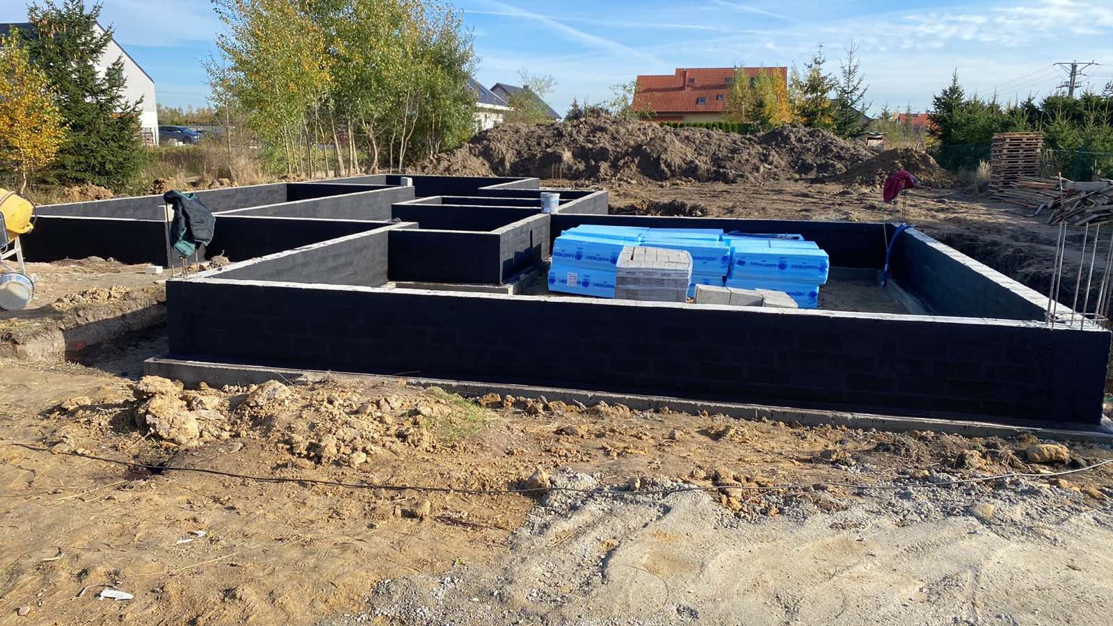 Fundamenty domu w trakcie budowy, zabezpieczone czarną izolacją, z widocznymi materiałami budowlanymi i ziemią w tle, w słoneczny dzień.