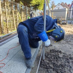 13th Welding Garage - Pracownik w niebieskim kombinezonie i masce spawalniczej łączy elementy metalowej konstrukcji na zewnątrz, widoczne są rękawice ochronne i przewód spawalniczy.