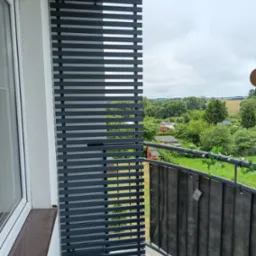 Szara, pozioma, lamelowa zabudowa balkonu z widokiem na zieleń, zamontowana obok okna. Balustrada z czarną przesłoną i dekoracją z bluszczu.