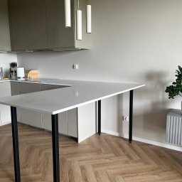 13th Welding Garage - Nowoczesna kuchnia z blatem w kształcie litery L, wsparta na czarnych, metalowych nogach. Parkiet w jodełkę, minimalistyczne oświetlenie i zabudowa kuchenna w tle.