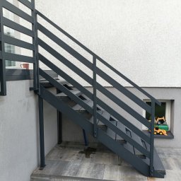 13th Welding Garage - Zewnętrzne schody metalowe z szarą, minimalistyczną balustradą, prowadzące do wejścia. W tle widoczna ściana budynku z oknem i zabawkami.