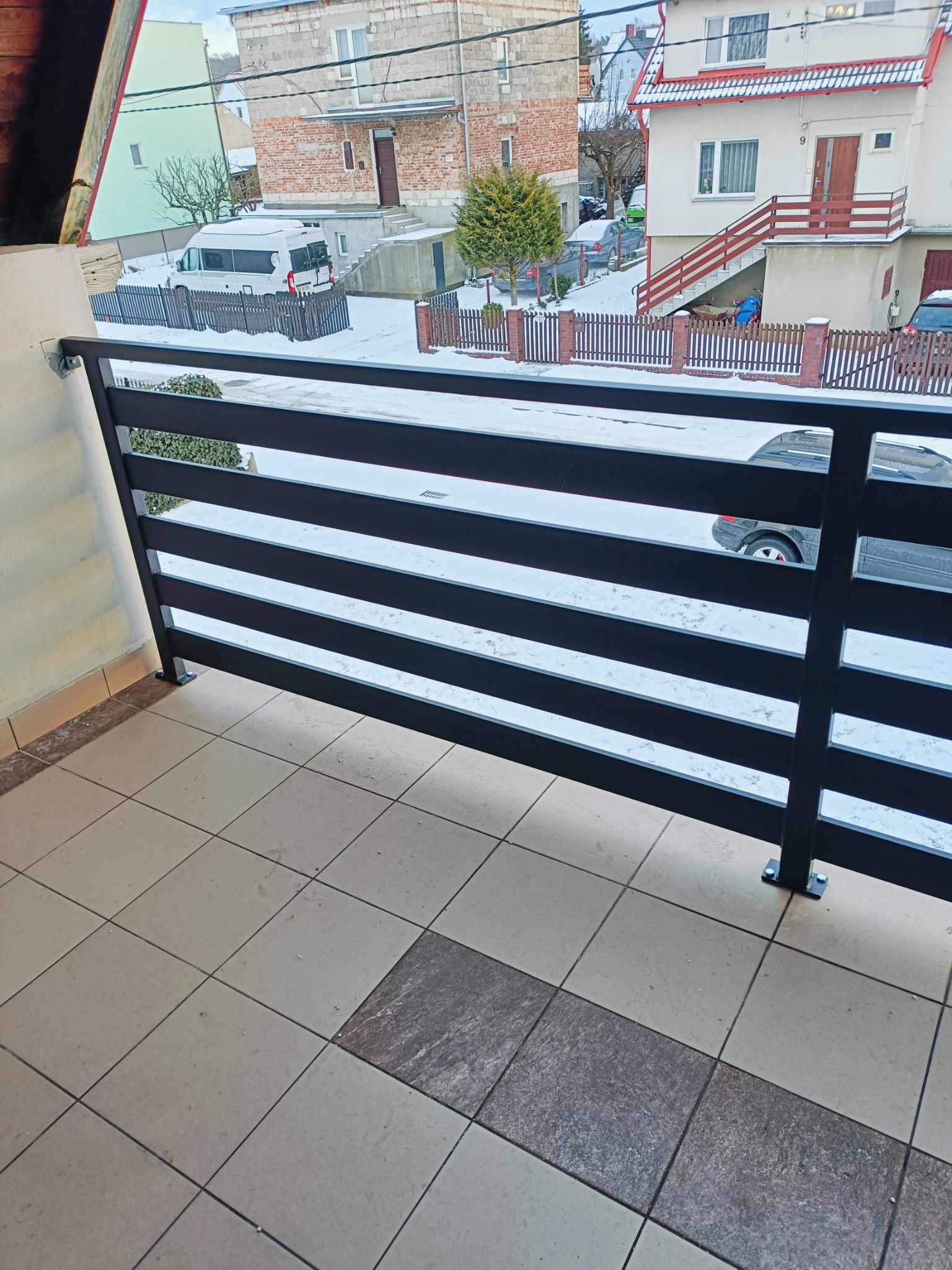 Czarna, pozioma balustrada metalowa na balkonie z widokiem na zasypane śniegiem podwórko i zabudowania w tle.