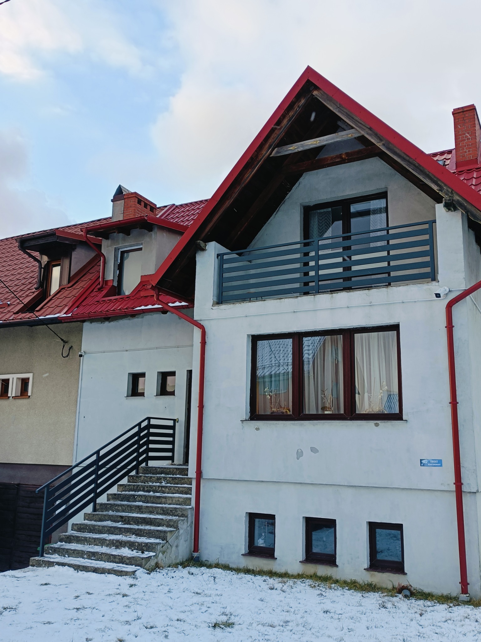 Dwupiętrowy dom z czerwonym dachem i czarną balustradą przy schodach wejściowych w zimowej scenerii.