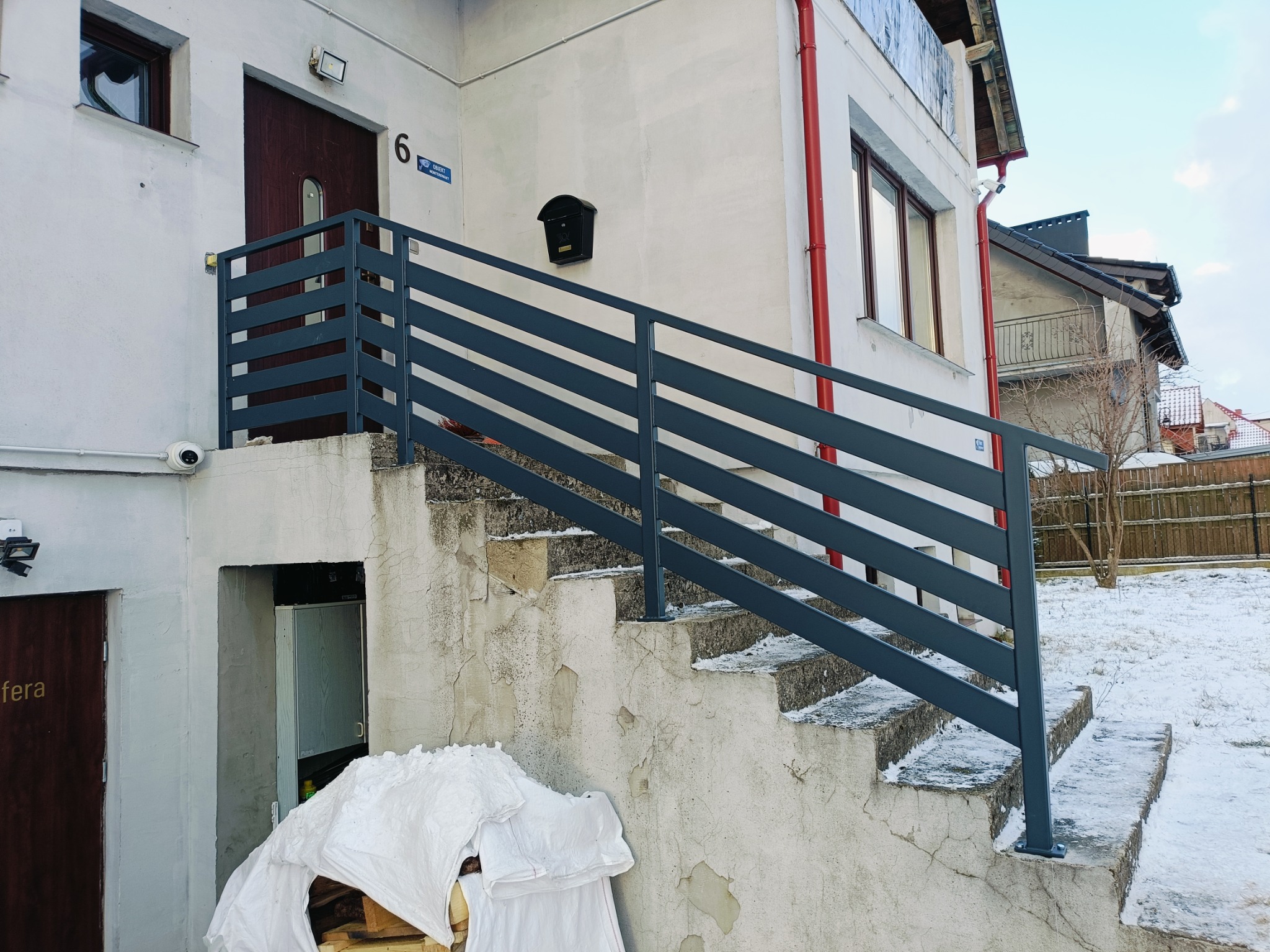 Zewnętrzne schody betonowe z nowoczesną, poziomą, szarą barierką przed budynkiem z numerem 6, widoczna skrzynka na listy i fragment ogrodu pokrytego śniegiem.
