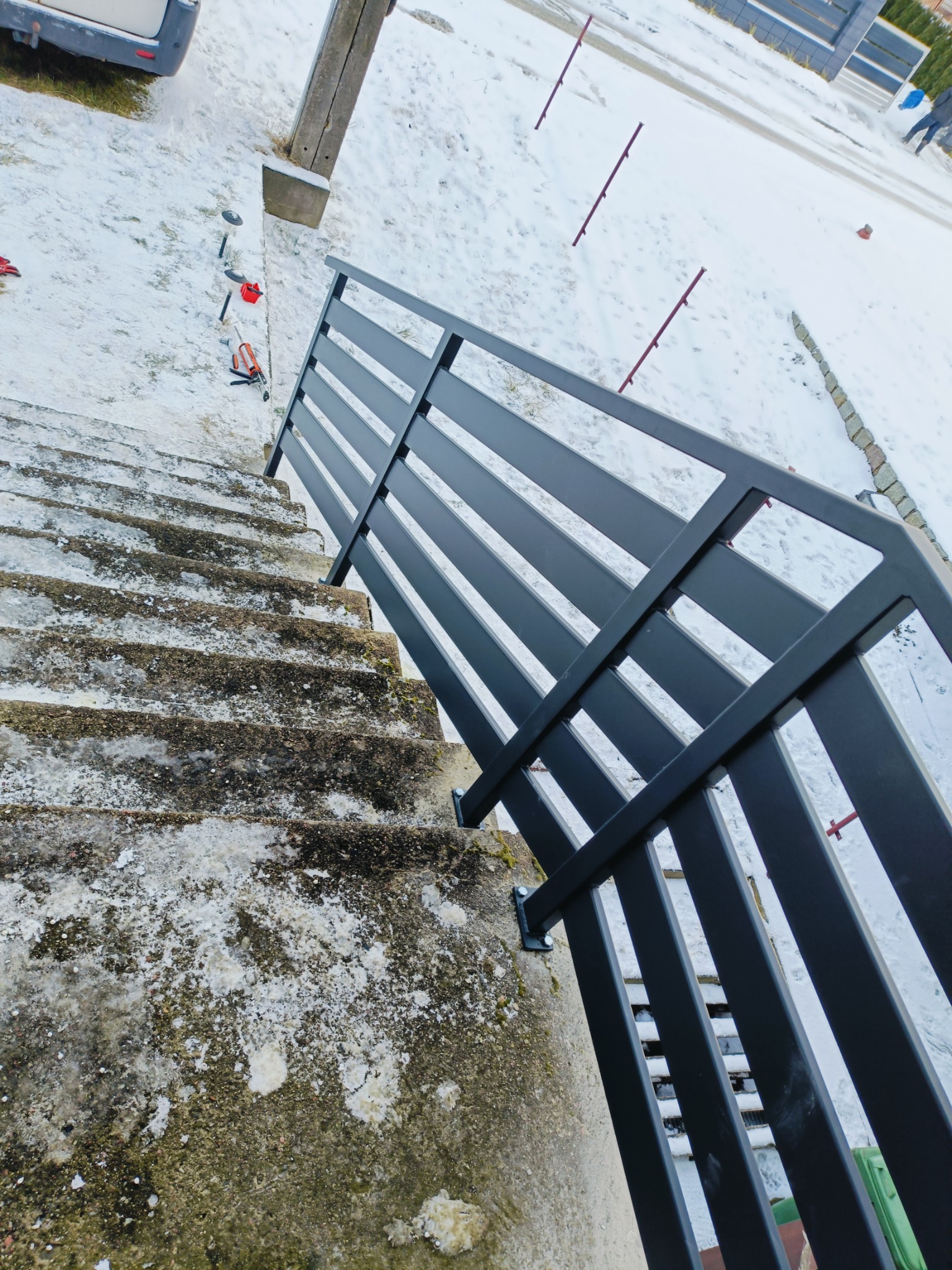 Częściowy widok nowo zamontowanej, czarnej, metalowej balustrady schodowej z profilami poziomymi, prowadzącej w dół po betonowych schodach zewnętrznych pokrytych śniegiem, z narzędziami montażowymi...