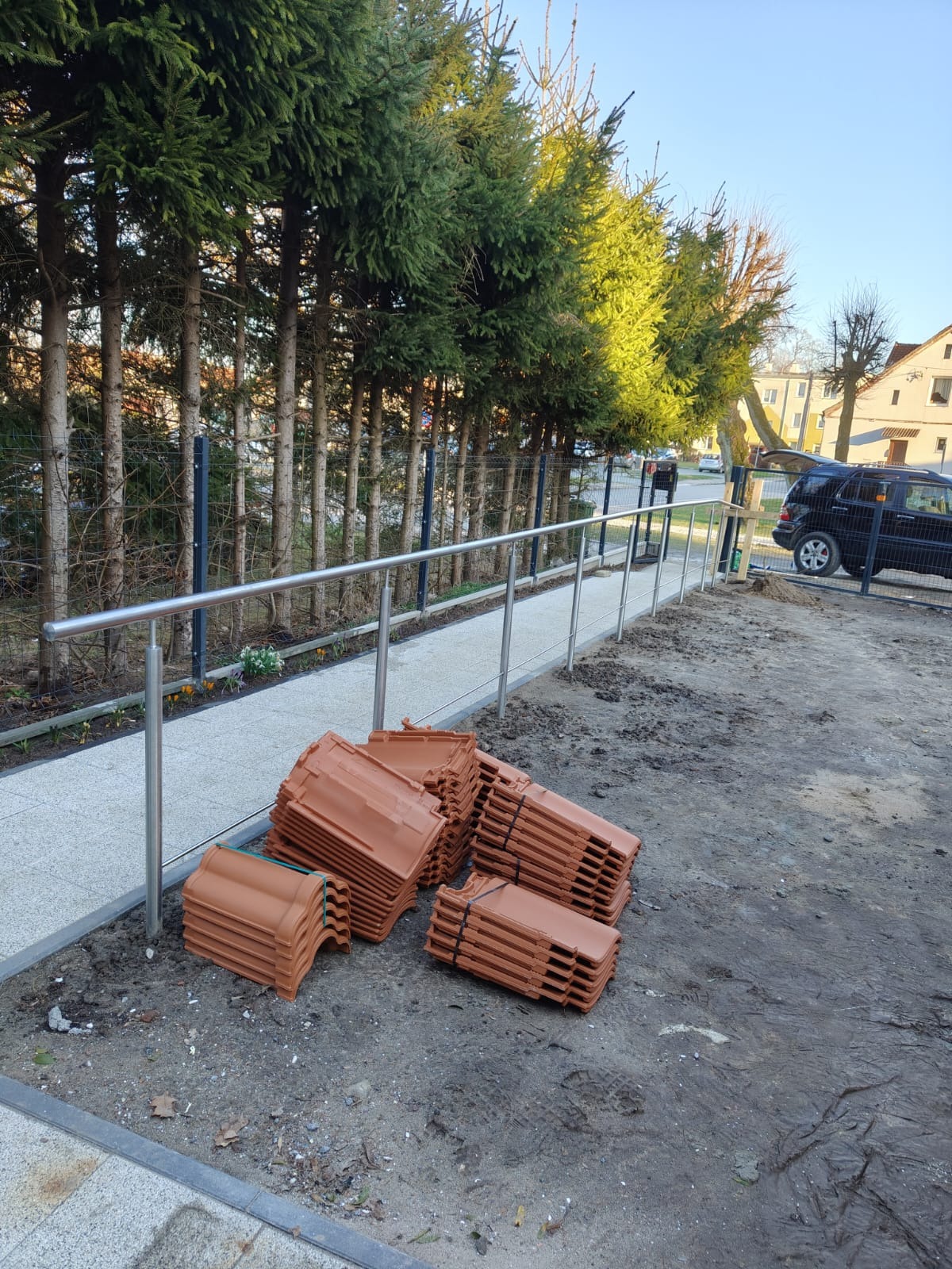 Stosy brązowych dachówek ceramicznych leżą na ziemi obok chodnika z nierdzewną balustradą, w tle rząd wysokich świerków i częściowo widoczny czarny samochód.