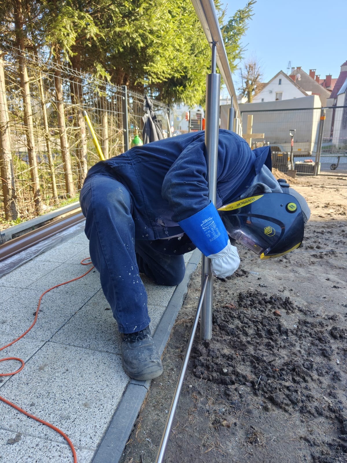 Pracownik w niebieskim kombinezonie i masce spawalniczej łączy elementy metalowej konstrukcji na zewnątrz, widoczne są rękawice ochronne i przewód spawalniczy.