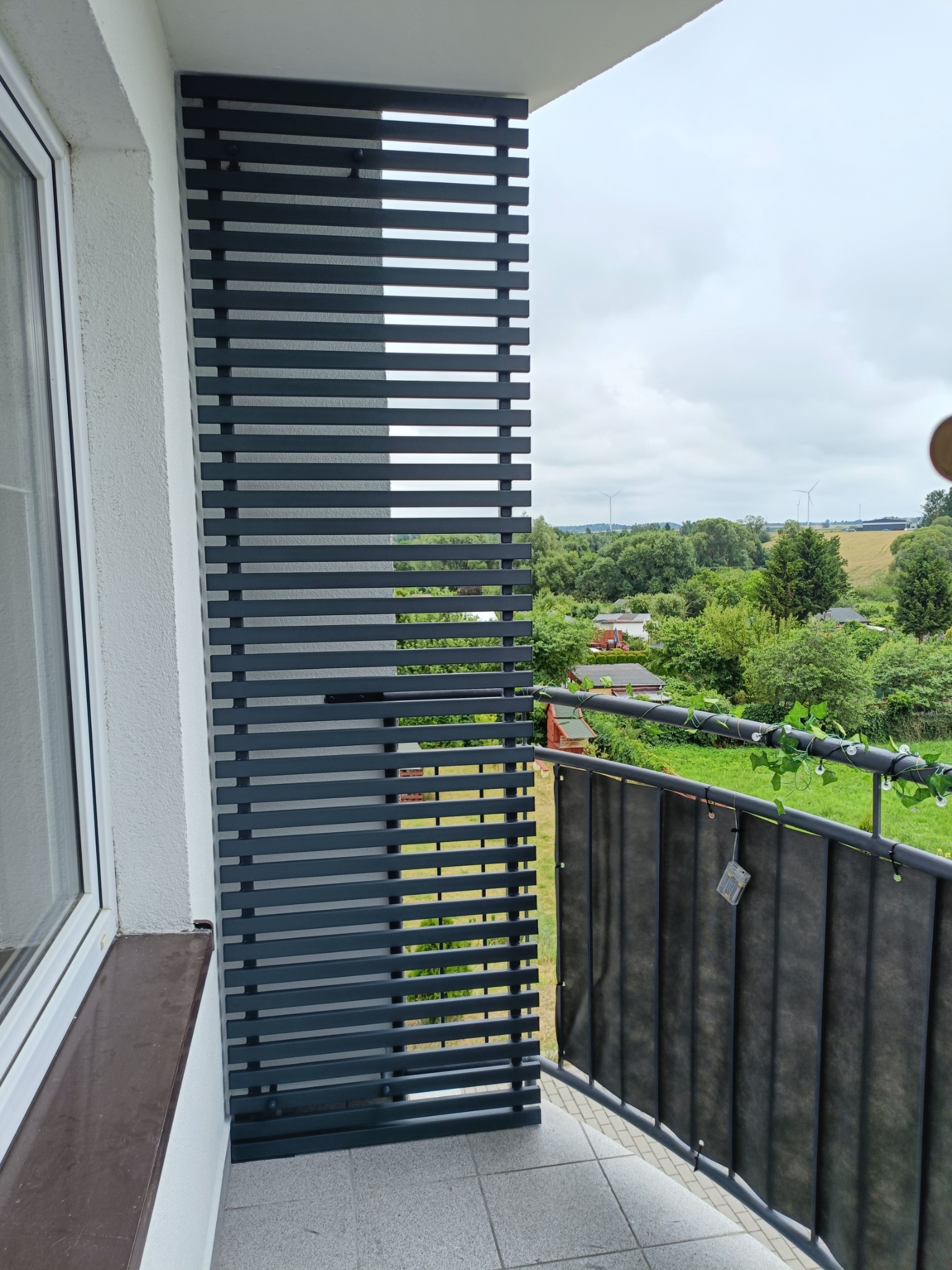 Szara, pozioma, lamelowa zabudowa balkonu z widokiem na zieleń, zamontowana obok okna. Balustrada z czarną przesłoną i dekoracją z bluszczu.