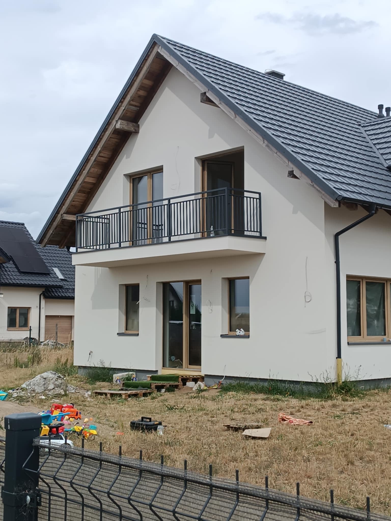 Nowoczesny dom z balkonem i czarną, metalową balustradą. Widoczne okna z drewnianymi ramami i szara dachówka. Na pierwszym planie fragment ogrodzenia panelowego.