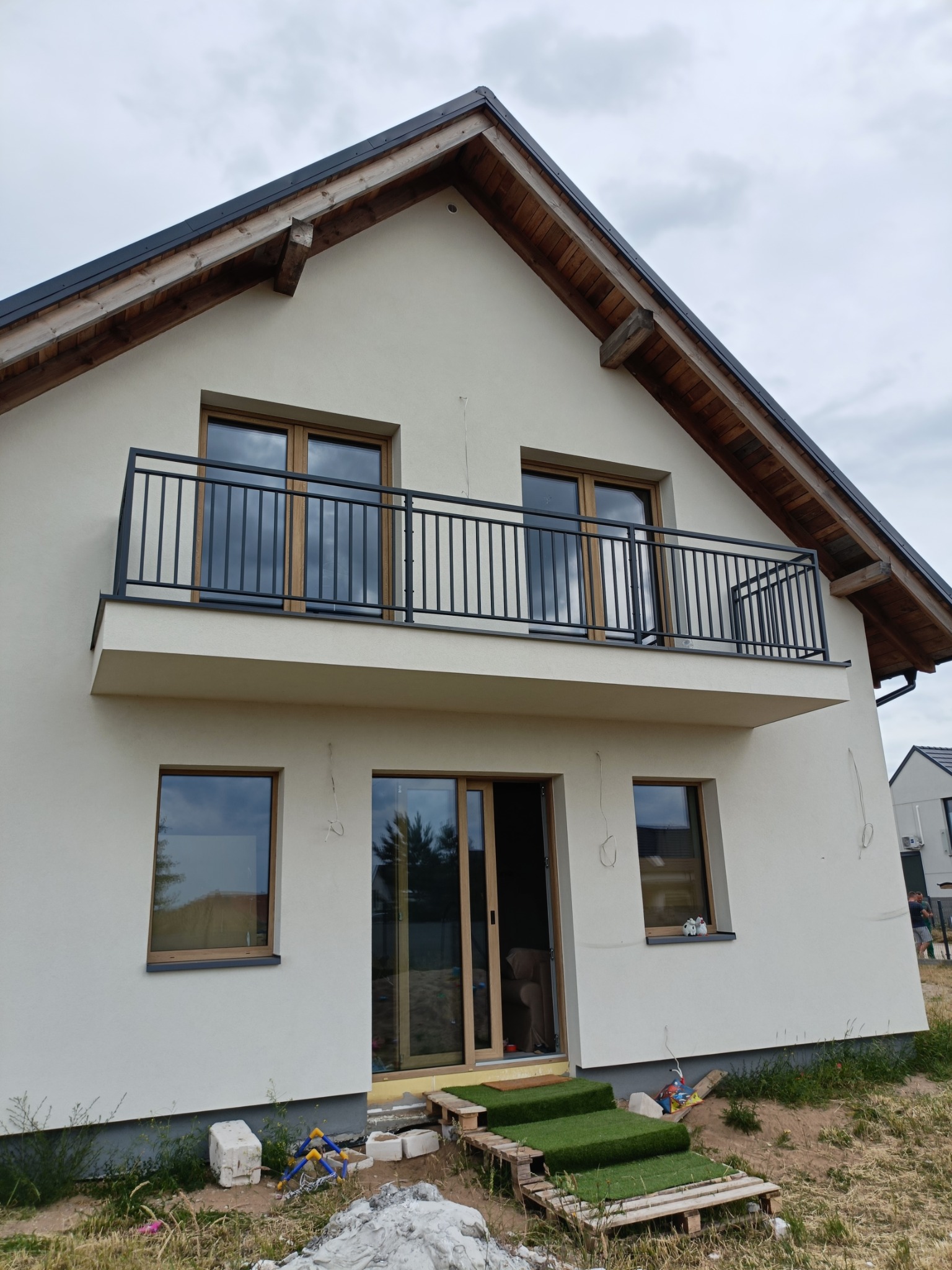 Dom z balkonem i czarną, metalową balustradą. Widoczne drewniane elementy dachu i tymczasowe schody z palet. Ujęcie z dołu, pochmurne niebo w tle.