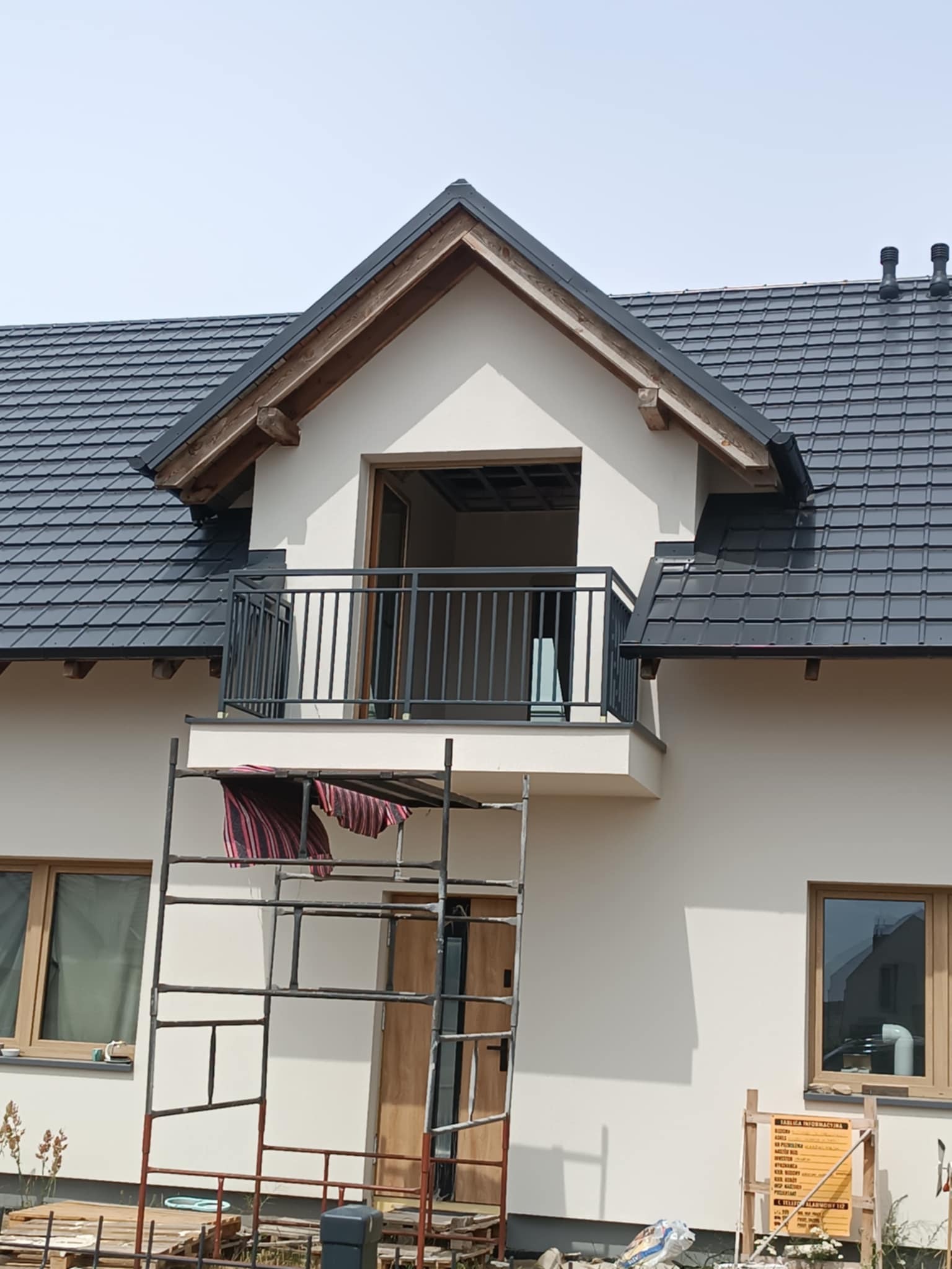 Wykończony balkon z czarną, metalową balustradą w nowym domu. Na pierwszym planie rusztowanie z rozwieszoną szmatką. Widoczne okna i dach z ciemnej dachówki.