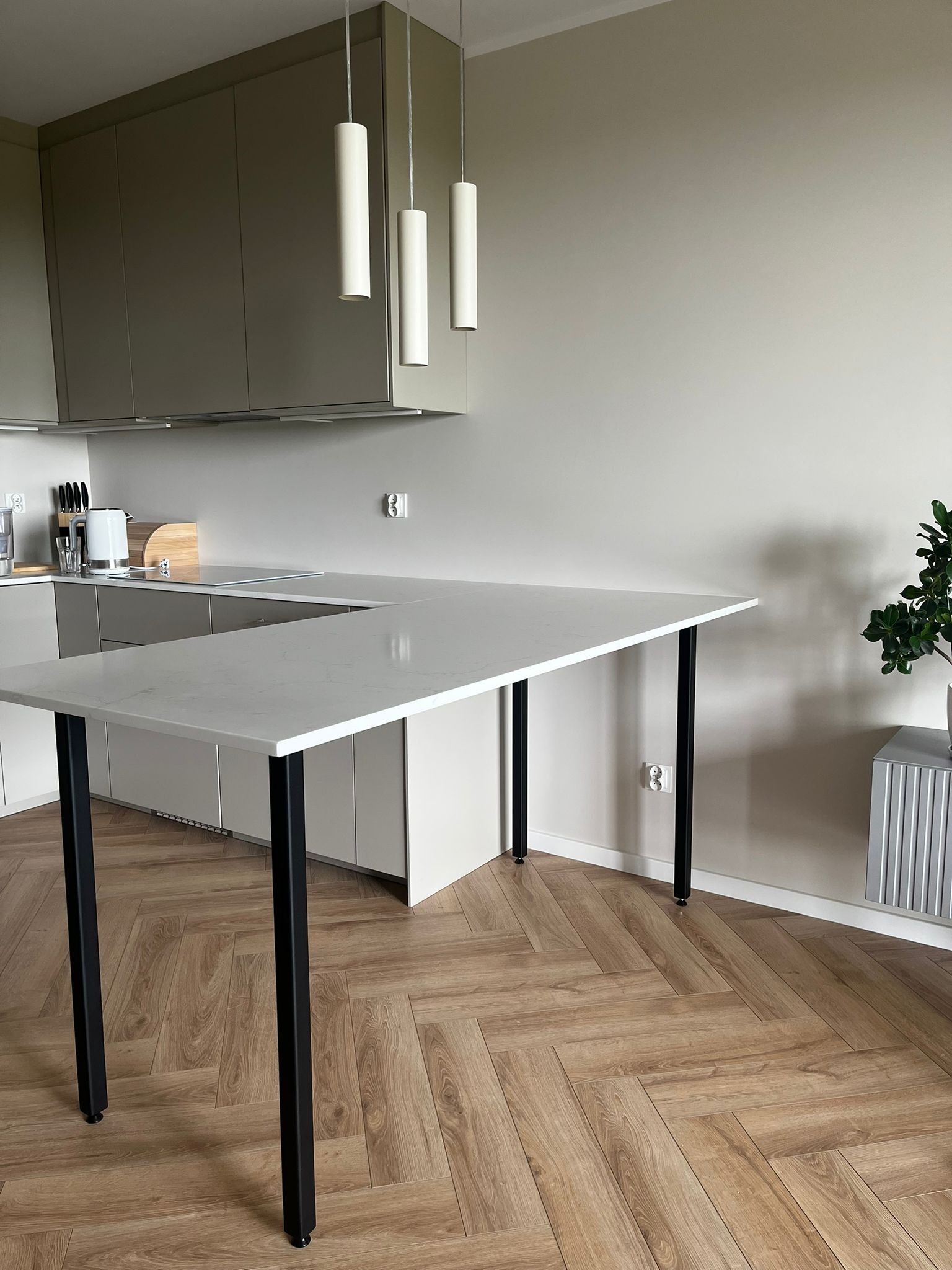 Nowoczesna kuchnia z blatem w kształcie litery L, wsparta na czarnych, metalowych nogach. Parkiet w jodełkę, minimalistyczne oświetlenie i zabudowa kuchenna w tle.