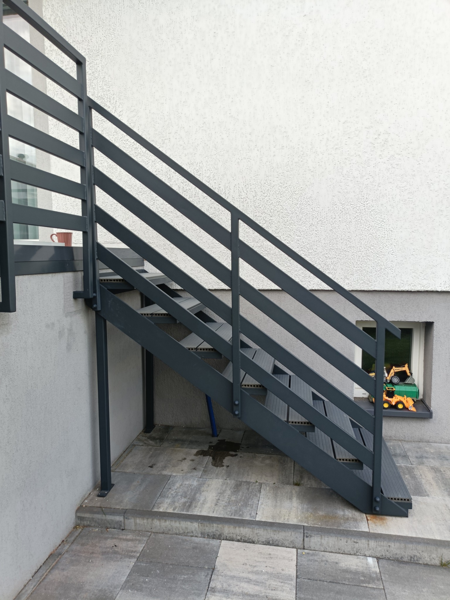 Zewnętrzne schody metalowe z szarą, minimalistyczną balustradą, prowadzące do wejścia. W tle widoczna ściana budynku z oknem i zabawkami.