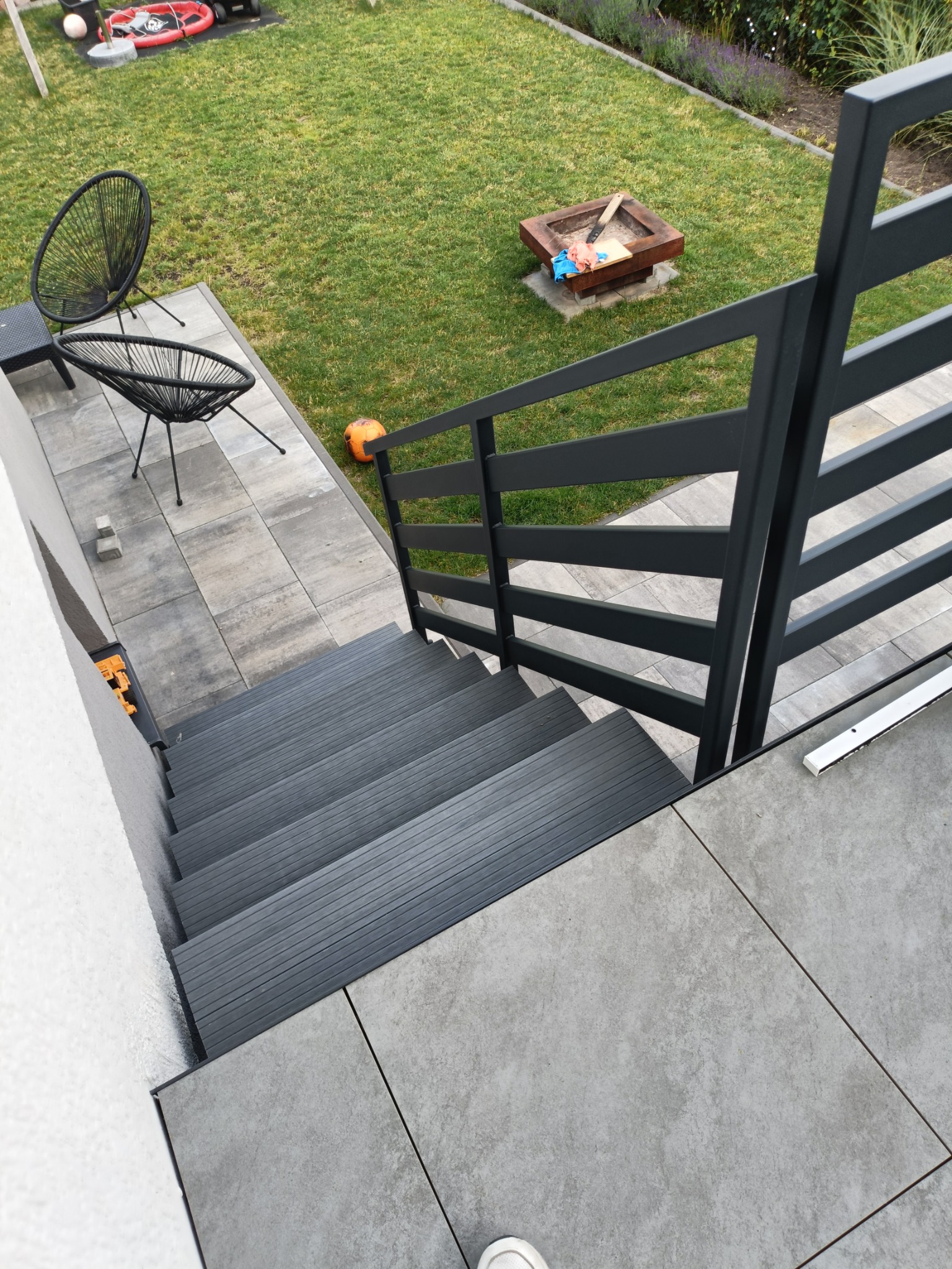 Nowoczesne schody zewnętrzne z szarymi stopniami i czarną, metalową balustradą na tle zadbanego trawnika z elementami rekreacyjnymi.