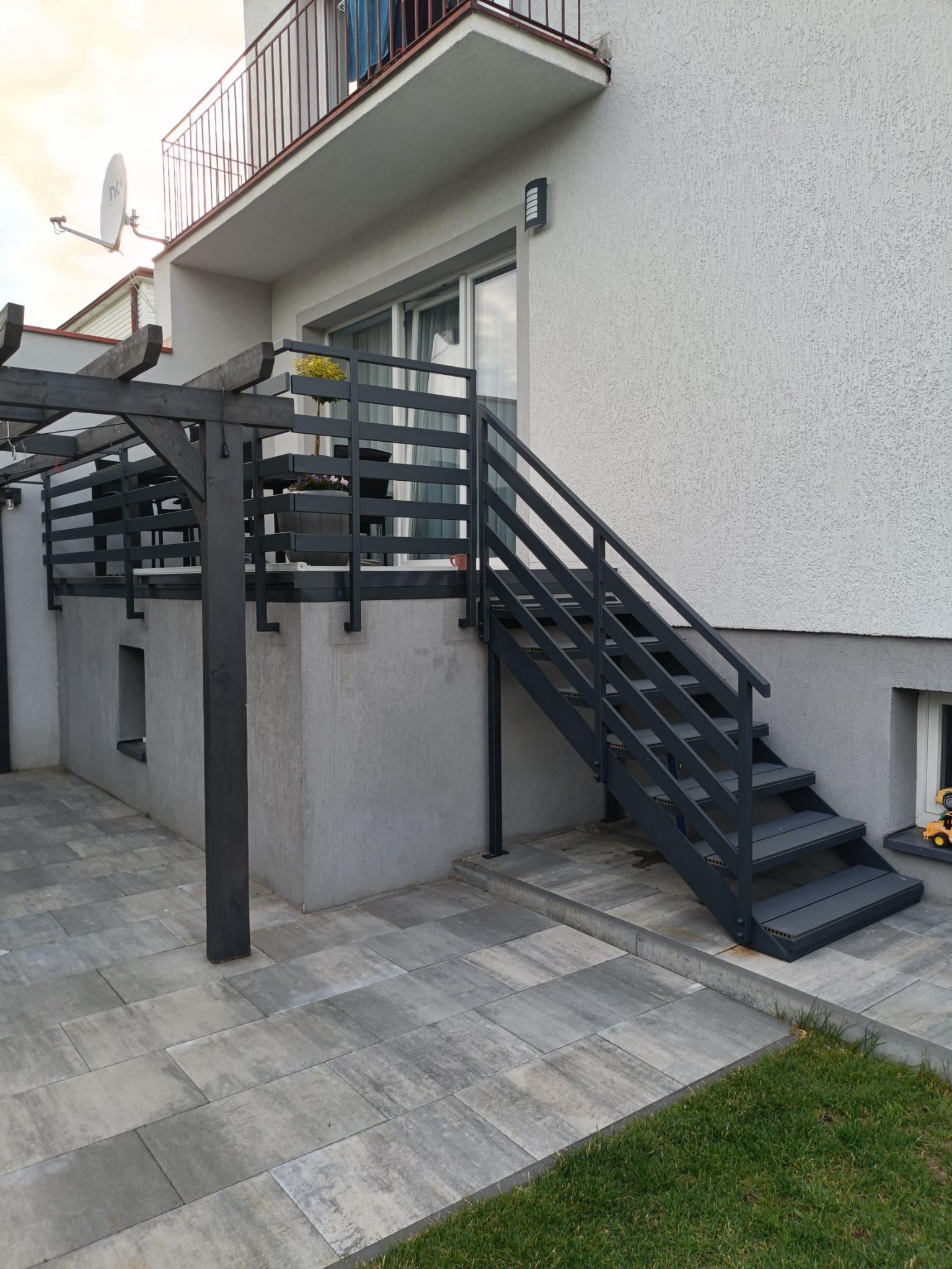 Zewnętrzne schody metalowe w grafitowym kolorze, prowadzące na balkon z pergolą. Nowoczesny wygląd, kostka brukowa i fragment trawnika. Elegancka architektura.