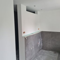 DoMpro- Remonty I Wykończenia wnętrz - Przygotowane do malowania ściany w pomieszczeniu z szarą glazurą, zabezpieczone folią malarską, widoczne puszki elektryczne i ślady farby na podłodze.