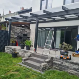 Zadaszenie tarasu z czarną konstrukcją i przezroczystym dachem, widoczne schody wejściowe, drabina i skrzynka z narzędziami na tle białego domu z dużymi oknami.