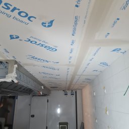DoMpro- Remonty I Wykończenia wnętrz - Wnętrze pomieszczenia w trakcie prac wykończeniowych, widoczne płyty gipsowe marek Glasroc i Rigips na suficie, wentylacja, białe płytki na ścianach z widocznymi punktami mocowania, podłoga...