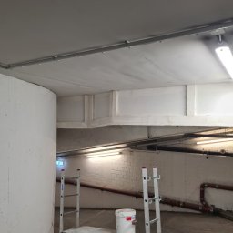 DoMpro- Remonty I Wykończenia wnętrz - Wnętrze w trakcie malowania: biała farba na suficie i ścianach, drabina z platformą, wiadro z farbą, widoczne rury pod sufitem, surowe oświetlenie, betonowa ściana.